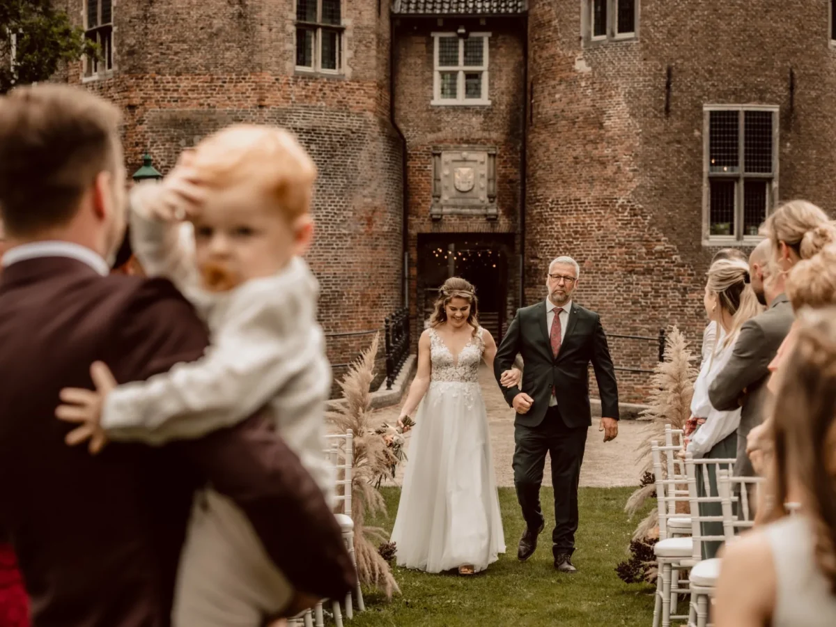 Bruid komt met vader aanlopen bij de ceremonie bij Kasteel Dussen
